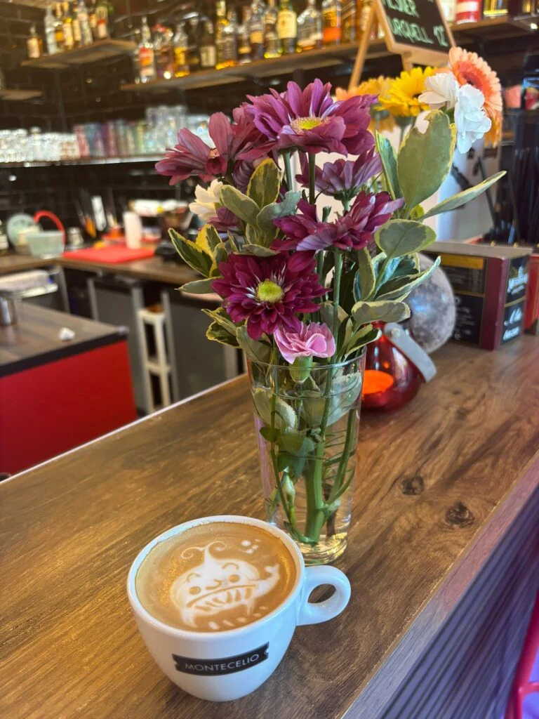 Cappuccino s veselým latte art v hrnku Montecelio na dřevěném baru s čerstvými květinami v kavárně Caffé GP Praha.